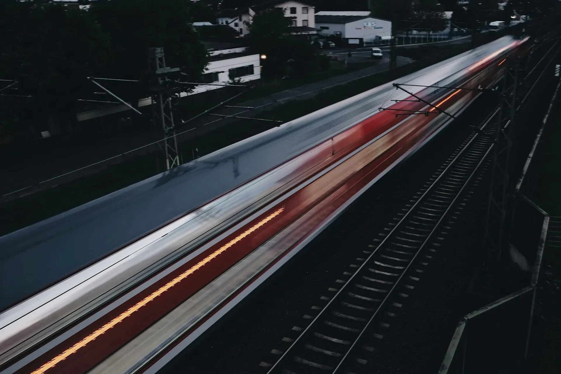 Trem passando em longa exposição ao entardecer, com rastros de luz vermelha e branca sobre os trilhos