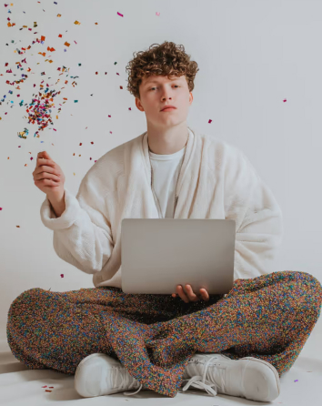 Curly-haired person in white robe sitting cross-legged with a laptop, tossing colorful confetti in the air.